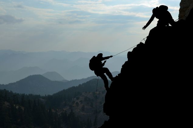 Imagem de uma pessoa escalando uma montanha e outra pessoa a esperando em cima, já no topo