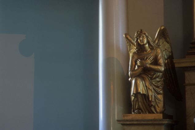 Imagem da estátua de um anjo ajoalhado, com as mãos no coração dentro de uma igreja
