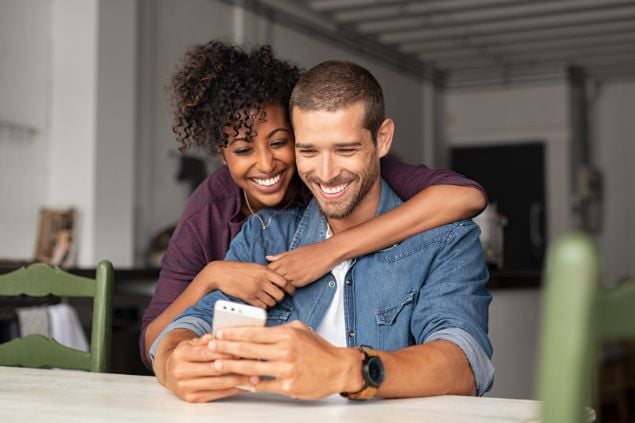 Imagem de um casal abraçado e vendo o celular na mesa da cozinha
