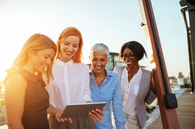 Quatro mulheres ao ar livre sorrindo, enquanto uma delas mostra algo em seu tablet