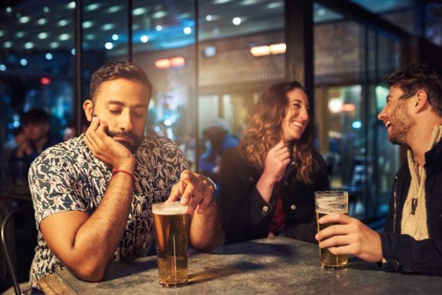 Homem em uma balada, sentado sozinho na bancada do bar e tomando cerveja, com expressão de tédio, enquanto um casal conversa ao seu lado