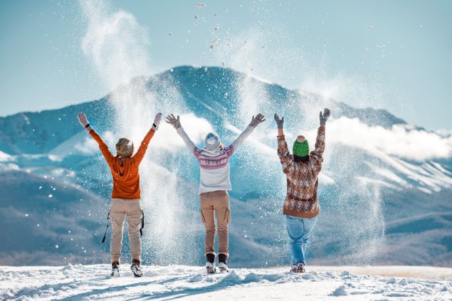 Três pessoas de costas, com os braços erguidos, brincando com a neve.
