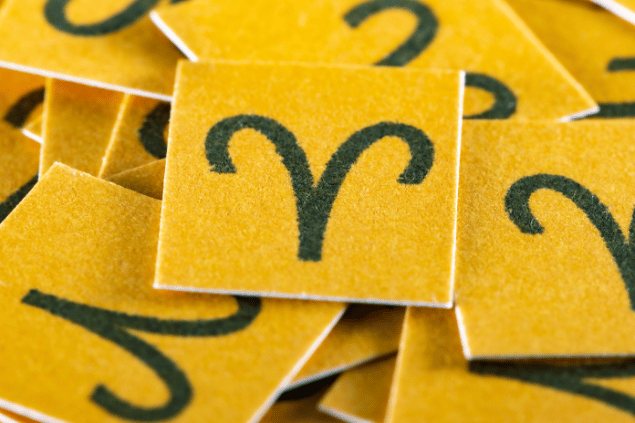 No foco da imagem, há vários pedaços de papel amarelo com o desenho do símbolo do signo de Áries.
