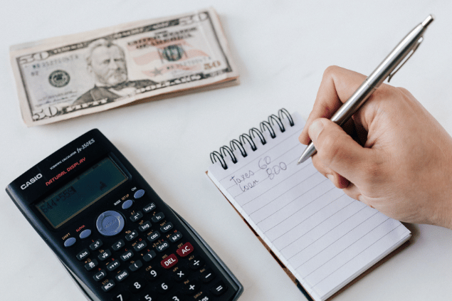 Mão escrevendo em um caderninho de anotações as despesas financeiras. Ao lado, notas de dólar e uma calculadoura.