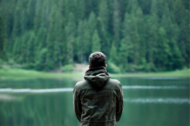 Homem de costas observando um lago e uma floresta ao fundo