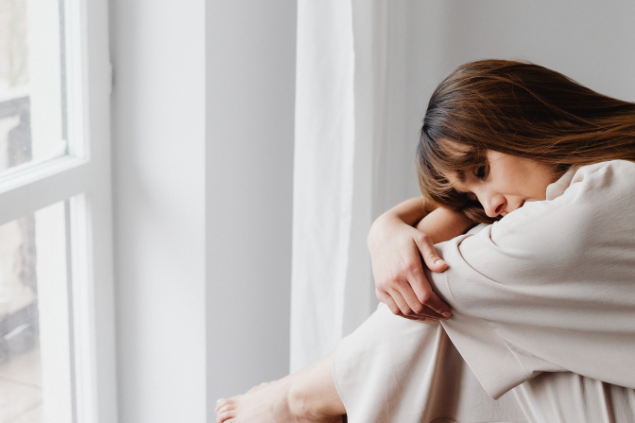 Mulher abraçando suas pernas enquanto está triste olhando pela janela