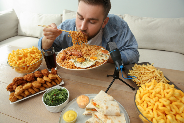 Homem com muita comida na boca numa mesa cheia de comida.