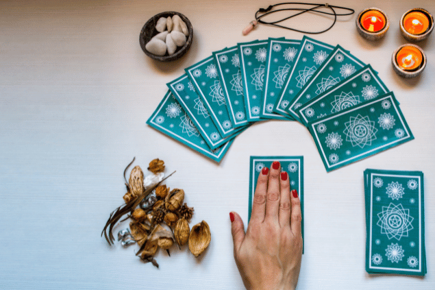 A mão de uma taróloga e uma tiragem de cartas de Tarot estão  sobre uma mesa e no foco da imagem. Ao lado, há velas, pedras e plantas.