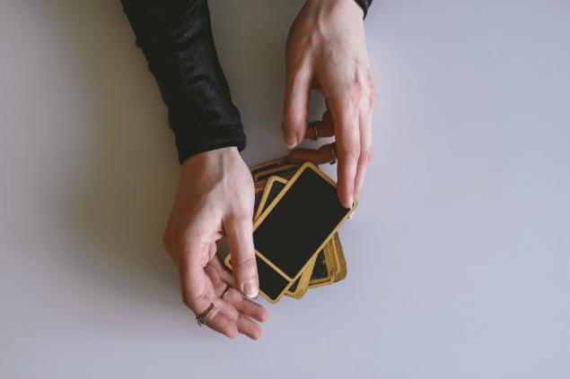 As mãos de uma taróloga segurando cartas de Tarot viradas para baixo estão em destaque na imagem.