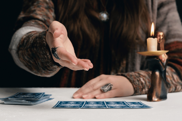 As mãos de uma taróloga  que estão em cima de uma mesa e perto de cartas de Tarot viradas para baixo estão em destaque na imagem.