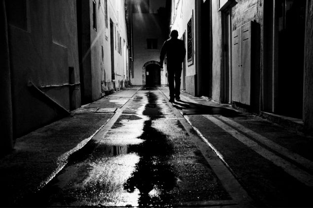 Imagem em preto e brando de um homem caminhando em uma rua deserta com o chão molhado, simbolizando o Exu da Meia-Noite.