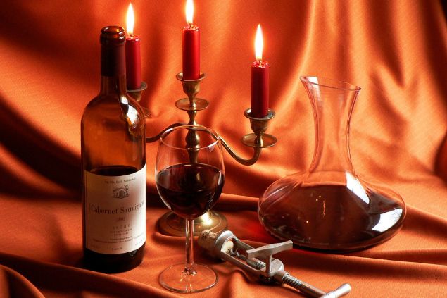 Imagem de uma mesa com uma toalha vermelha. Sobre ela, uma garrafa de vinho tindo, taça, abridor e um castiçal com velas vermelhas acesas, simbolizando uma oferenda para o Exu da Meia-Noite.