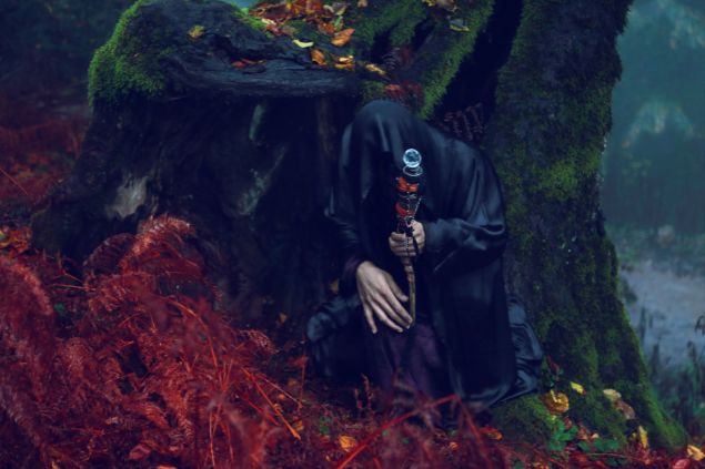 Imagem de um homem usando uma capa preta, encostado em um tronco com lodo, simbolizando o Exu Capa Preta.