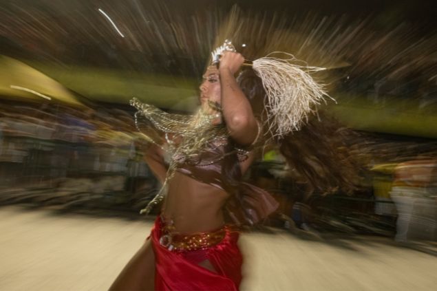Imagem de uma mulher, dançando em um terreira de  umbanda. Ela usa uma saia vermelha e um top marrom com colares dourados, simbolizando a pombagira Maria Mulambo.