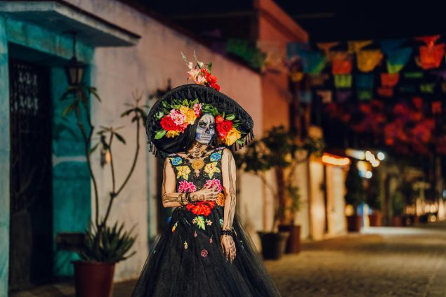 Imagem de uma mulher usando uma pintura de caveira no rosto, além de um chapéu e um vestido longo preto, decorado com flores coloridas.