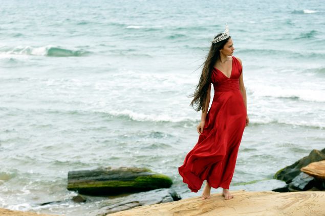 Imagem de uma mulher na beira da praia, usando um vestido vermelho e uma coroa na cabeça, simbolizando a pombagira Rainha.