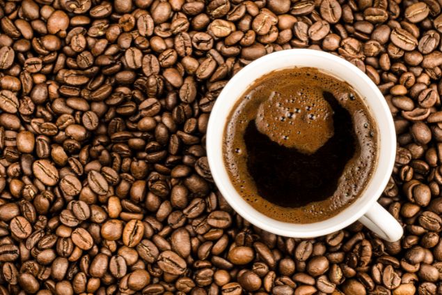 Imagem de um xícara branca contendo café preto sobre vários grãos da bebida. Trata-se de um item utilizado nas oferendas ao preto velho Pai Joaquim de Angola.