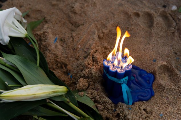 Imagem de um punhado de velas acesas na cor azul depositada na areia da praia. Ao  lado, um ramalhete de flores brancas, simbolizando o ebó.