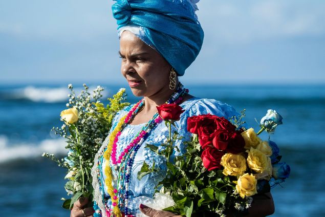 Imagem de uma guia espiritual feminina, usando um turbante e um vestido azul, com várias guias coloridas, segurando muitas rosas amarelas e vermelhas. Ela está na praia e simboliza a Iyalorixá no Candomblé.
