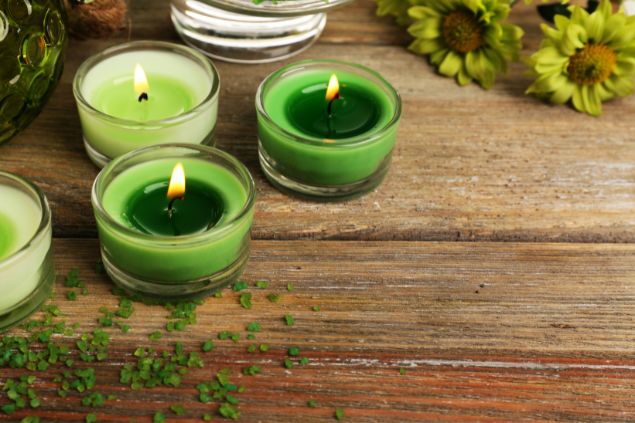 Pixelshot / Canva Imagem de várias velas acesas na cor verde, dispostas sobre uma mesa de madeira, decorada com algumas folhas verdes, simbolizando uma das oferendas para Zé Pereira.
