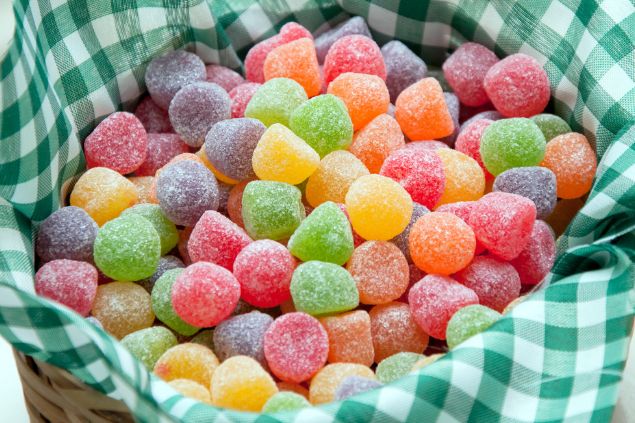 JuFagundes / Getty Images / Canva Imagem de uma cesta com tecido xadrez nas cores verde e branco, contendo balas de jujuba coloridas, uma das oferendas para o Erê Joãozinho da Umbanda.