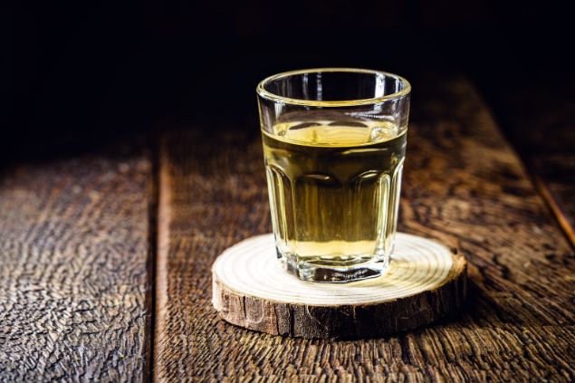 RHJ / Getty Images / Canva Imagem de um copo com dose de pinga sobre uma mesa de madeira, simbolizando uma das oferendas para o Exu Tatá Caveira.