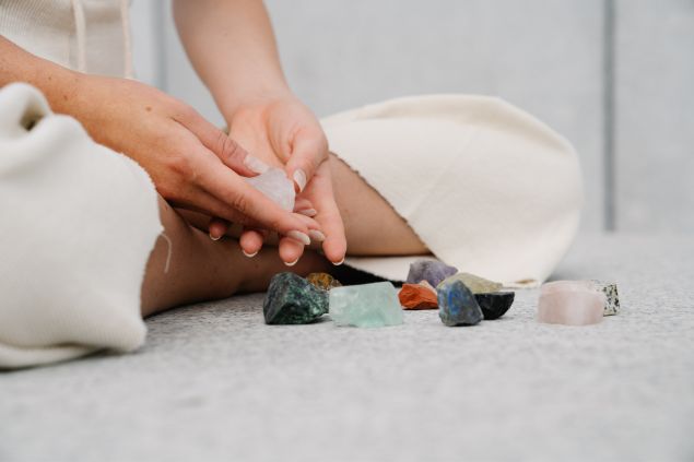 Kevin Malik / Pexels / Canva Imagem de uma reikiana sentada sobre um tapete branco, manipulando alguns cristais e pedras para renovar suas energias para a prática do reiki.