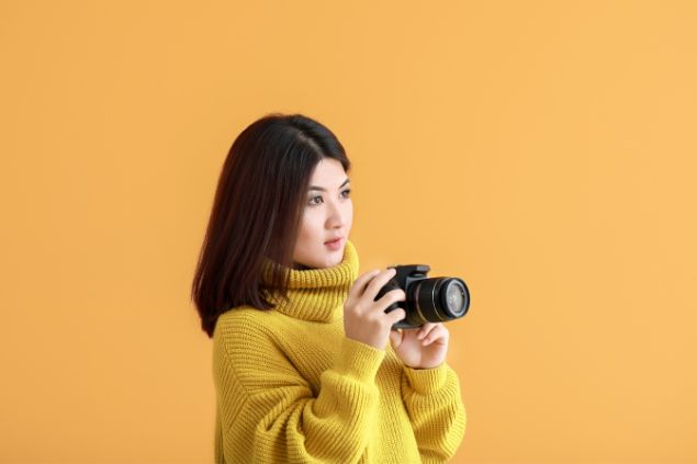 Pixelshot / Canva Imagem de fundo amarelo e em destaque uma mulher fotógrafa segurando uma câmera em mãos.