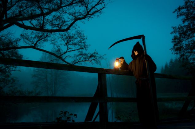 Shaiith / Getty Images / Canva Imagem de uma noite nebulosa e em destaque um homem com a roupa da morte segurando em uma das mãos uma foice e na outra uma lanterna. Ele está parado em uma ponte velha