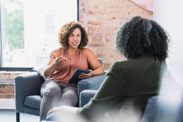SDI Productions / Getty Images Signature / Canva Imagem de duas pessoas sentadas em um sofá em uma sala de estar. Elas estão trocando conselhos e uma delas é a líder emocional.