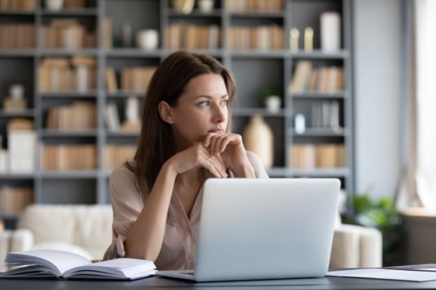 Fizkes / Getty Images / Canva Imagem de uma mulher designer. Ela está de frente com um computador, olhando pela janela e pensando no conceito criativo de suas peças.