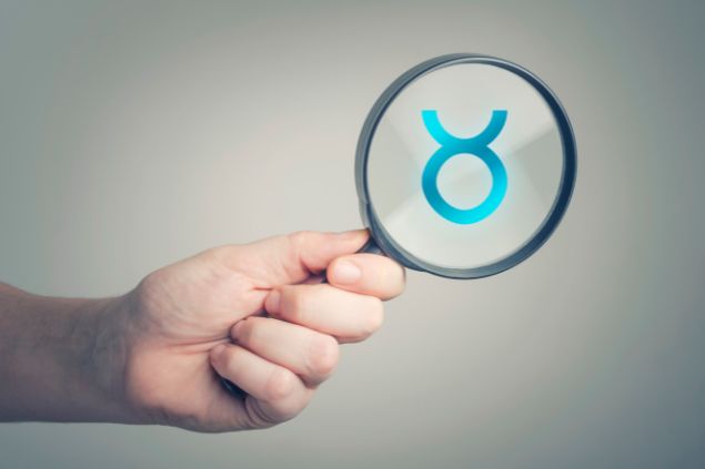 Natasaadzic / Getty Images / Canva Imagem do logo do signo de touro em destaque em uma lupa, sendo segurada pela mão de uma pessoa.