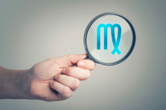 Natasaadzic / Getty Images / Canva Imagem do logo do signo de virgem em destaque em uma lupa, sendo segurada pela mão de uma pessoa.