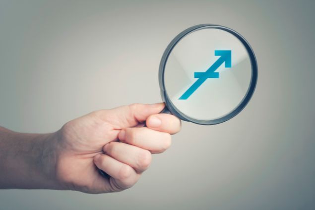 Natasaadzic / Getty Images / Canva Imagem do logo do signo de sagitário em destaque em uma lupa, sendo segurada pela mão de uma pessoa.