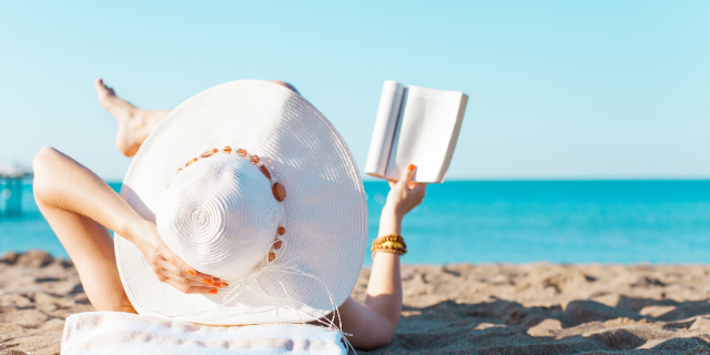 Mulher em praia, deitada sobre a areia e lendo um livro. Suas pernas estão cruzadas. Em uma das mãos, ela segura o próprio chapéu.