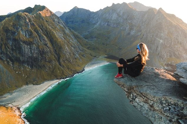 Mulher sentada na beira de um precipício. Abaixo dela, águas azuis e uma praia, com montanhas ao redor.