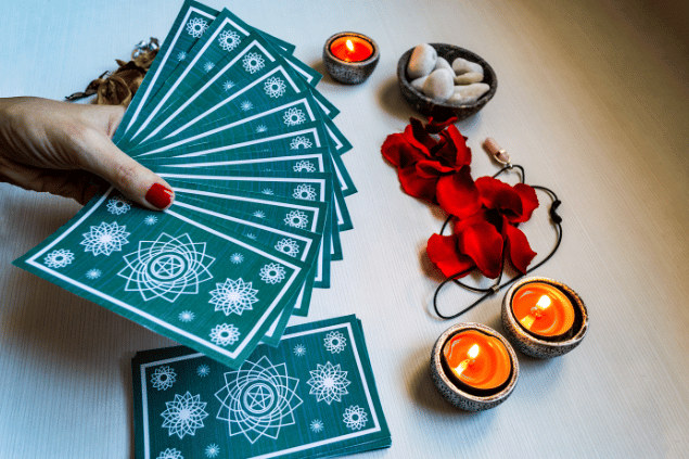 Baralho de tarot em uma mesa de madeira, com velas, flores e cristais. A mão de uma mulher segura algumas cartas. 