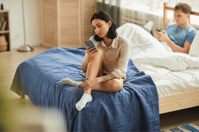 Mulher mexendo no celular distante do seu namorado, que está deitado na cama. 