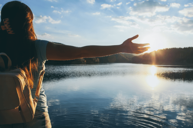 Mulher com os braços abertos diante um lago e uma vista ensolarada. 