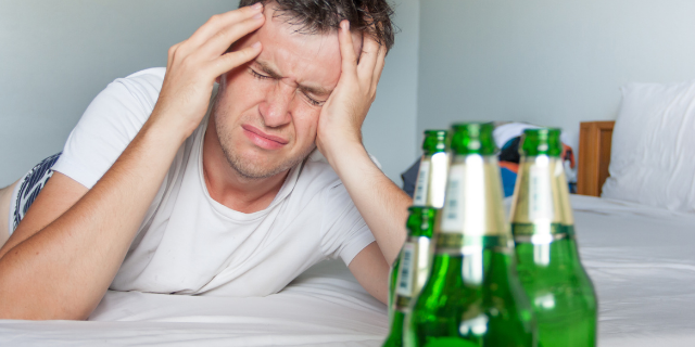 Homem deitado de bruços pressiona as têmporas em sinal de dor de cabeça. Em sua frente, há garrafas de cerveja.