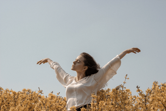Maksim Goncharenok / Pexels / Canva Uma mulher está com os braços abertos em um ambiente de natureza. Ela levanta a cabeça, fecha os olhos e sente a brisa do vento.