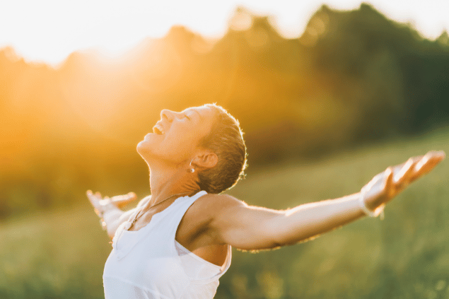Microgen / Getty Images / Canva Uma mulher está com os braços abertos em um ambiente de natureza. Ela levanta a cabeça e sente os raios de sol na pele.