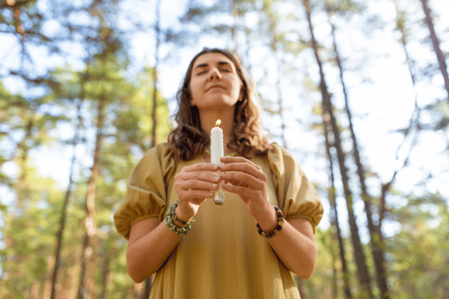 Uma mulher está segurando uma vela acesa em uma floresta. Ela fecha os olhos e sente o ambiente.