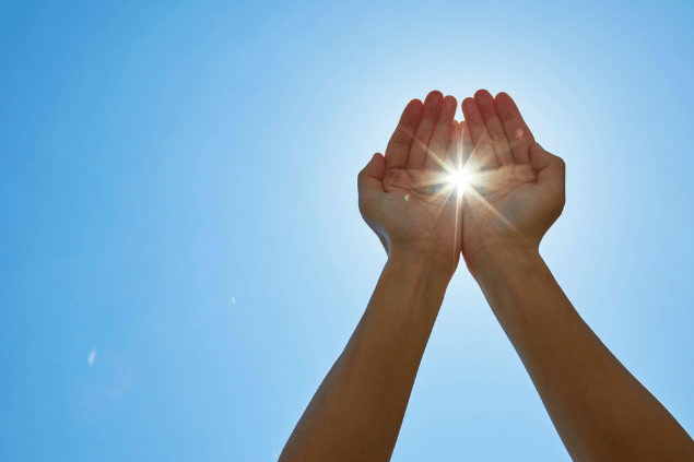 Aflo Images / アフロ(Aflo)/ Canva Uma pessoa está com os braços levantados e as mãos juntas em frente da luz do sol. Ao fundo, há o céu azul.