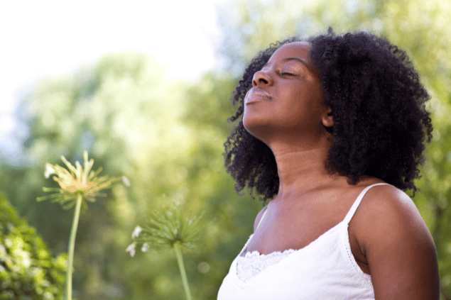 Uma mulher está em um ambiente de natureza. Ela levanta a cabeça, fecha os olhos e sente os raios de sol na pele.