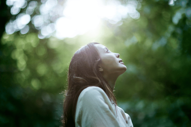 Aflo Images / アフロ(Aflo)/ Canva Uma mulher está em uma floresta. Ela levanta a cabeça e sente os raios de sol na pele.