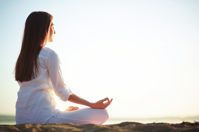 Pressmaster / Canva Uma mulher está sentada e meditando em uma praia. Ela sente os raios de sol na pele.