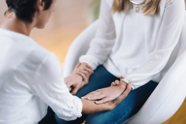 Duas mulheres estão de frente de uma para a outra. Elas seguram as mãos, usam roupas brancas e meditam.