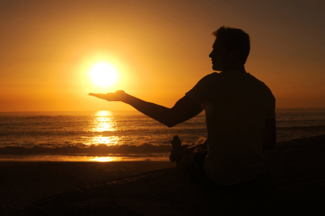 PeopleImages / Getty Images Signature / Canva Um homem está sentado de frente para o mar em uma praia. Ele observa e levanta uma mão em direção ao pôr do sol.