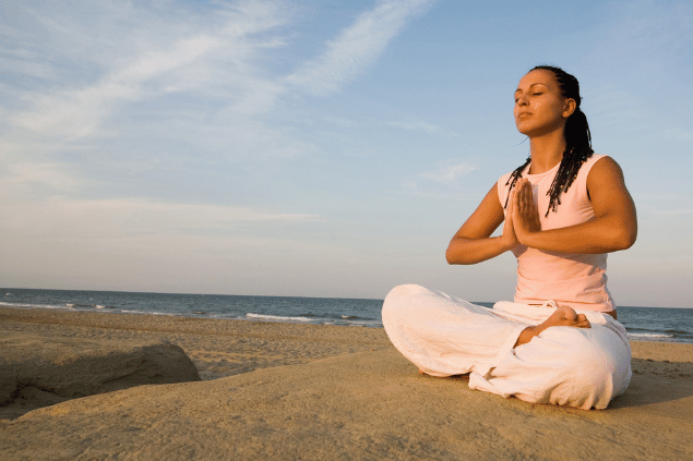 Aflo Images / アフロ(Aflo)/ Canva Uma mulher está sentada e meditando na praia sob a luz do sol.